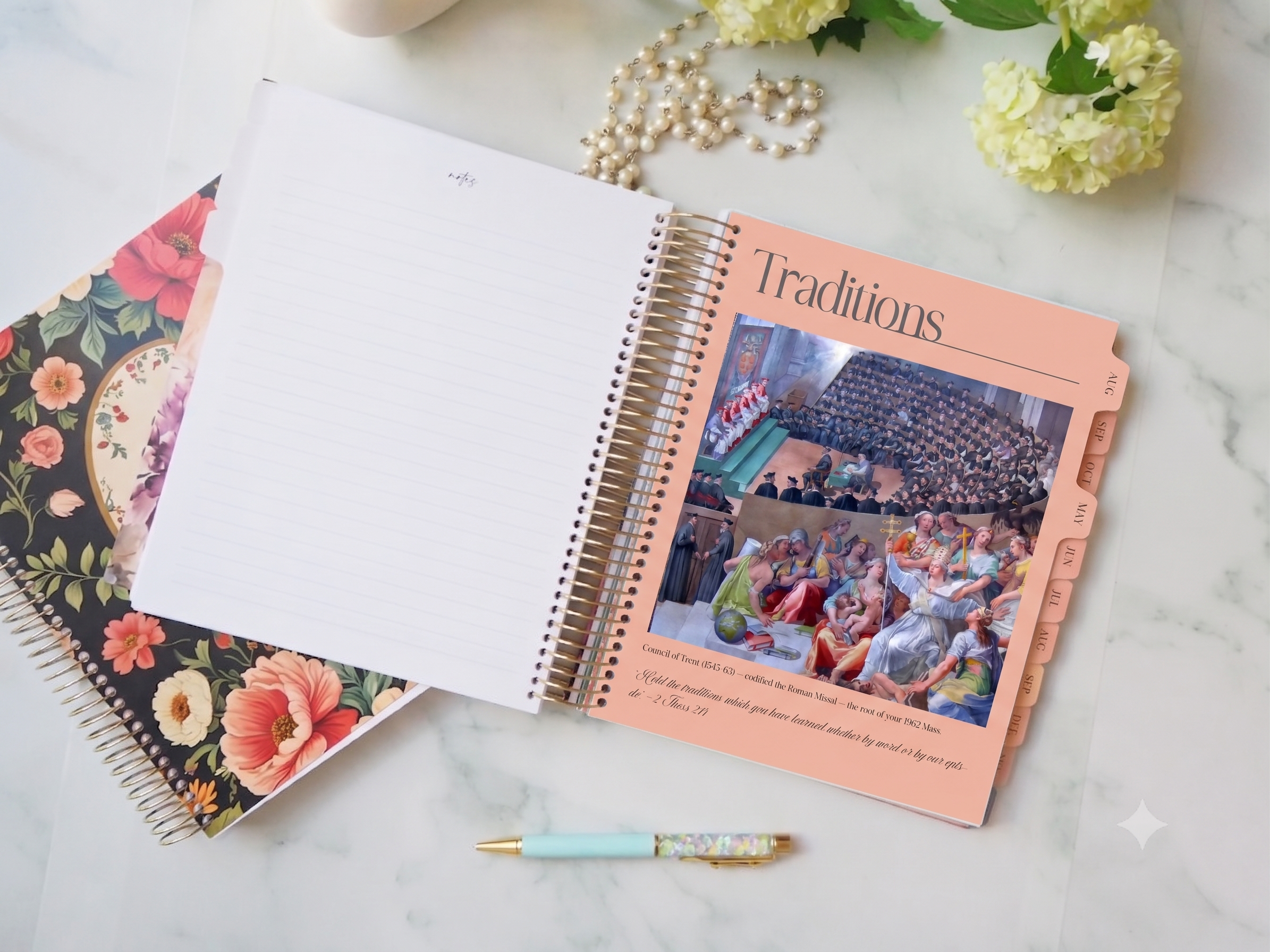 An open spiral-bound notebook or planner lies on a light marble surface, showing a blank white page on the left and a peach-colored right page titled "Traditions" with a printed wedding scene and tabbed pages. A floral-patterned notebook, a pastel pen, and decorative flowers and pearls surround it in a styled flat-lay.