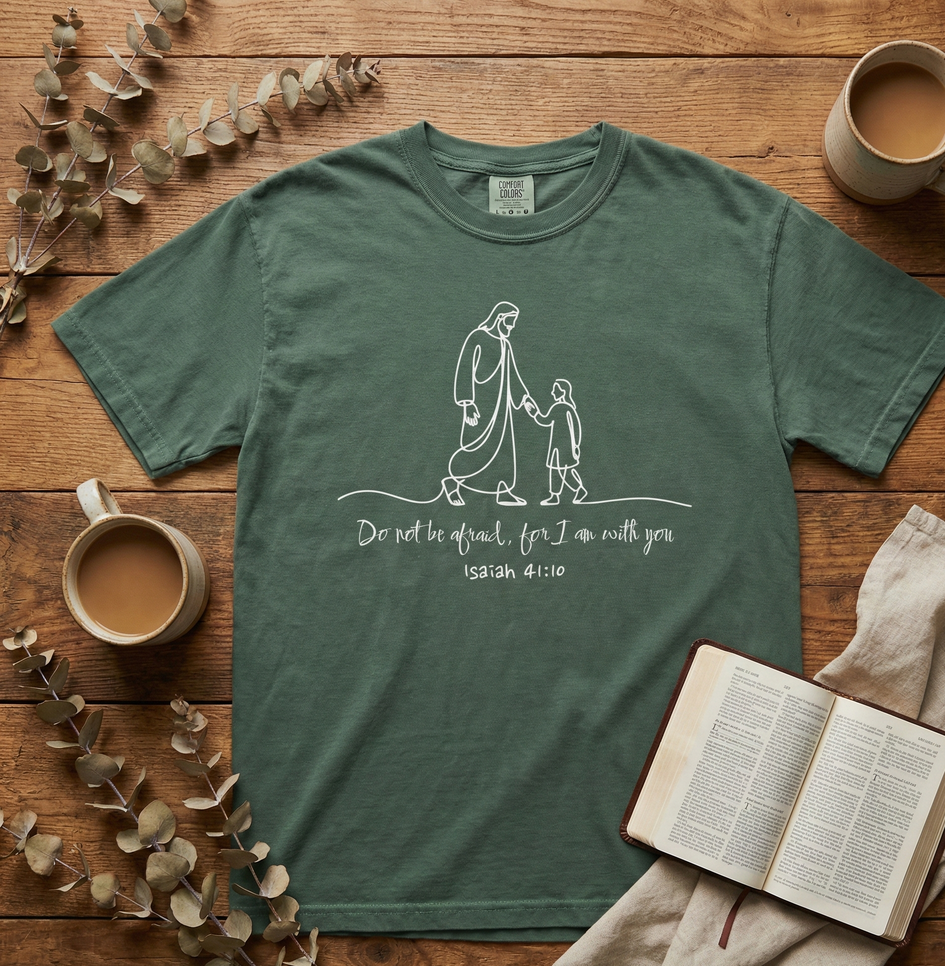 Green t-shirt with a religious design on a wooden surface with coffee cups and an open book.